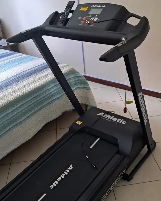 Esteira Athletic Speedy preta montada ao lado de uma cama em um quarto residencial.